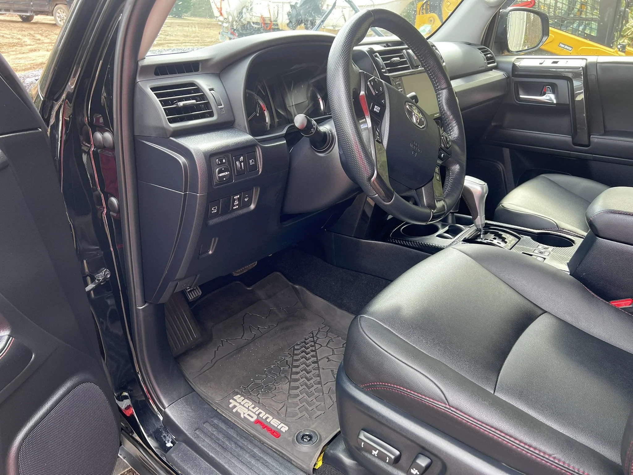Inside the driver's side of a black Toyota Land Cruiser Prado, showing the dashboard, steering wheel, gear shift, and black leather seats with red stitching, along with a custom all-weather floor mat.