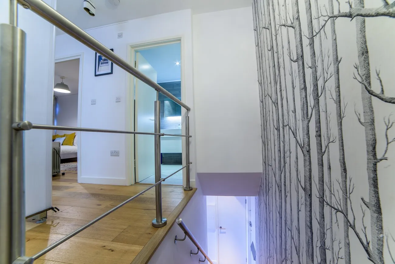Interior view of a hallway with a wooden floor and white walls, leading to rooms with doors open, one room visible with yellow pillows on a white sofa, a wall with black and white tree branch wallpaper, and a stainless steel railing along the staircase.