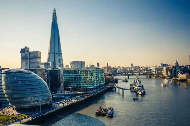 City skyline along a river with the Shard skyscraper, City Hall, and modern buildings in London, UK.