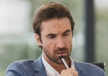 A man with dark hair and a beard, wearing a blue suit, appears to be thinking and is holding a pen to his mouth.
