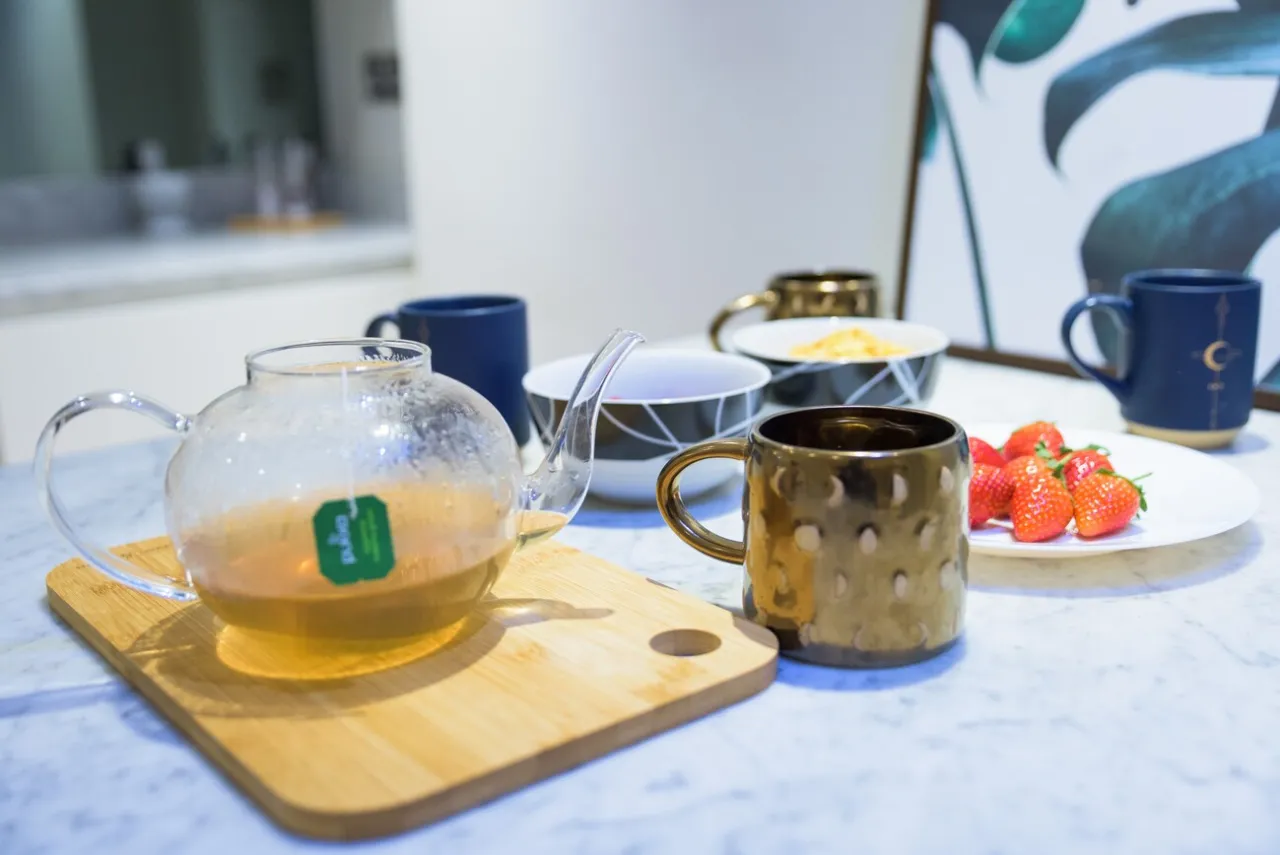 Tea set with a glass teapot, cups, a white plate with strawberries, and bowls with breakfast food on a marble countertop.
