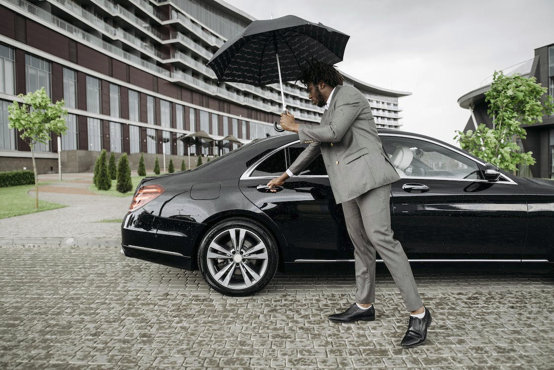 A man in a gray suit holding an umbrella and opening the front passenger door of a black luxury car on a paved area, with a modern building and small trees in the background, on a rainy day.