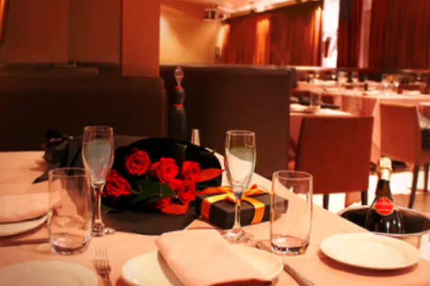 A restaurant table set for a celebration with a bouquet of red roses, a gift box wrapped with a ribbon, champagne bottles in a bucket, and multiple glasses.