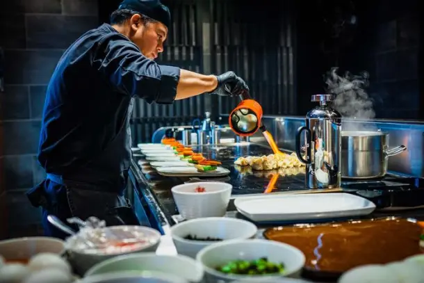 Chef preparing and pouring sauce over food in a professional kitchen with various dishes and cooking equipment on the counter.