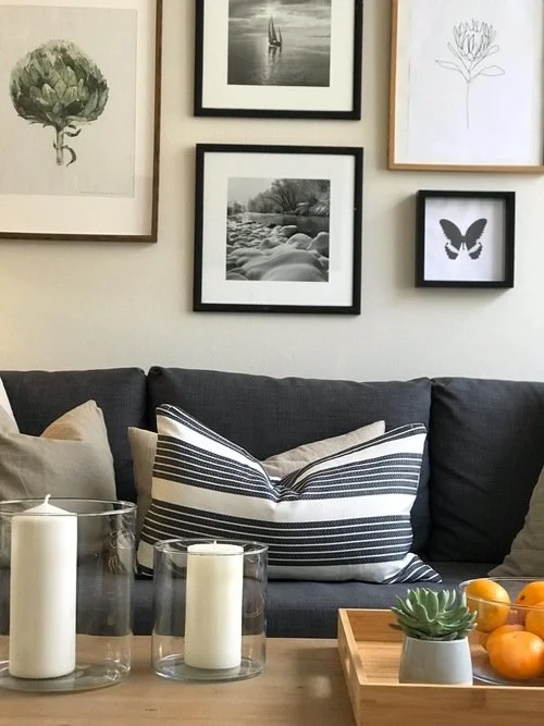Living room with a gray couch, striped pillows, candles in glass holders, and framed wall art including a black and white photo of a sailboat, a river scene, botanical sketches, and a butterfly illustration.