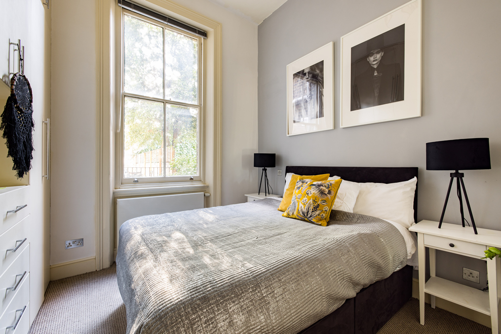 A cozy bedroom with a large window letting in natural light, a bed with a dark headboard, white and yellow decorative pillows, and two black table lamps on white side tables. Gray walls adorned with black-and-white framed photographs.