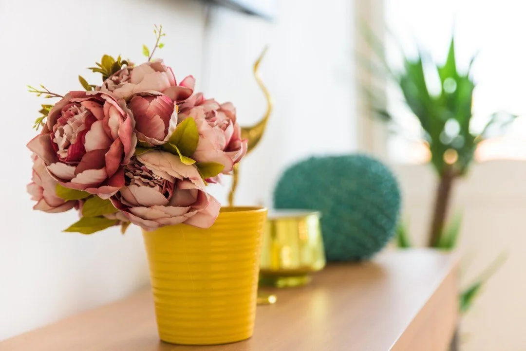 A pink peony flower in a yellow pot, with decorative gold objects and green plants in the background.
