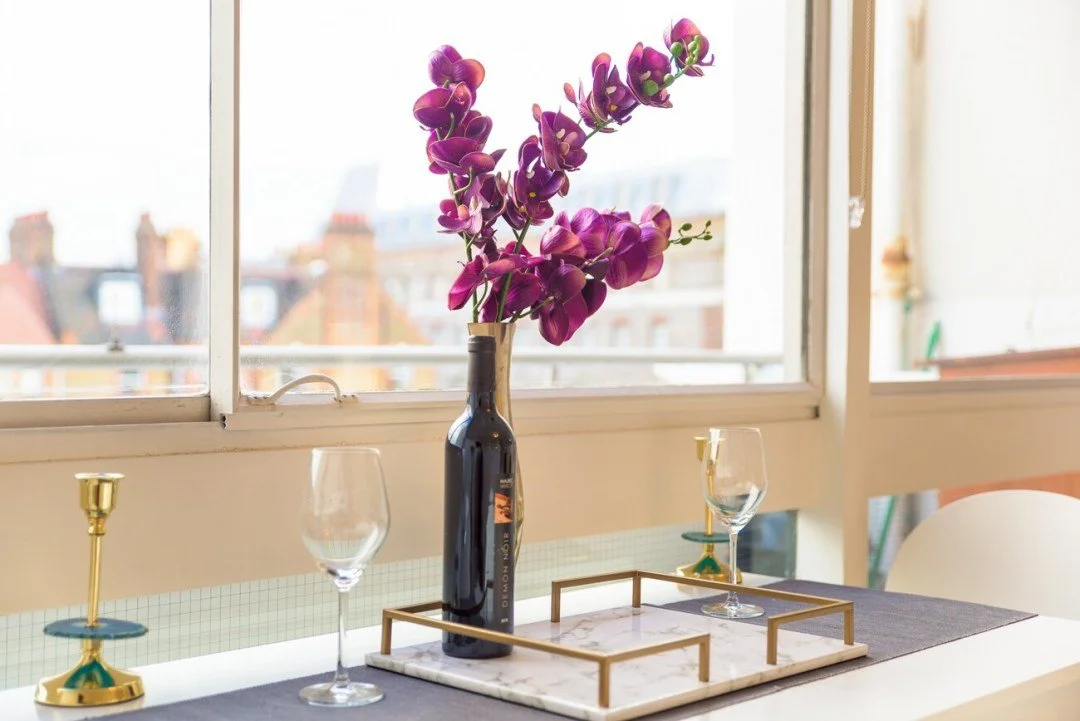 Decorative table setting with a black vase holding purple orchids, two wine glasses, and gold candlesticks on a gray table runner near a window with a city view.