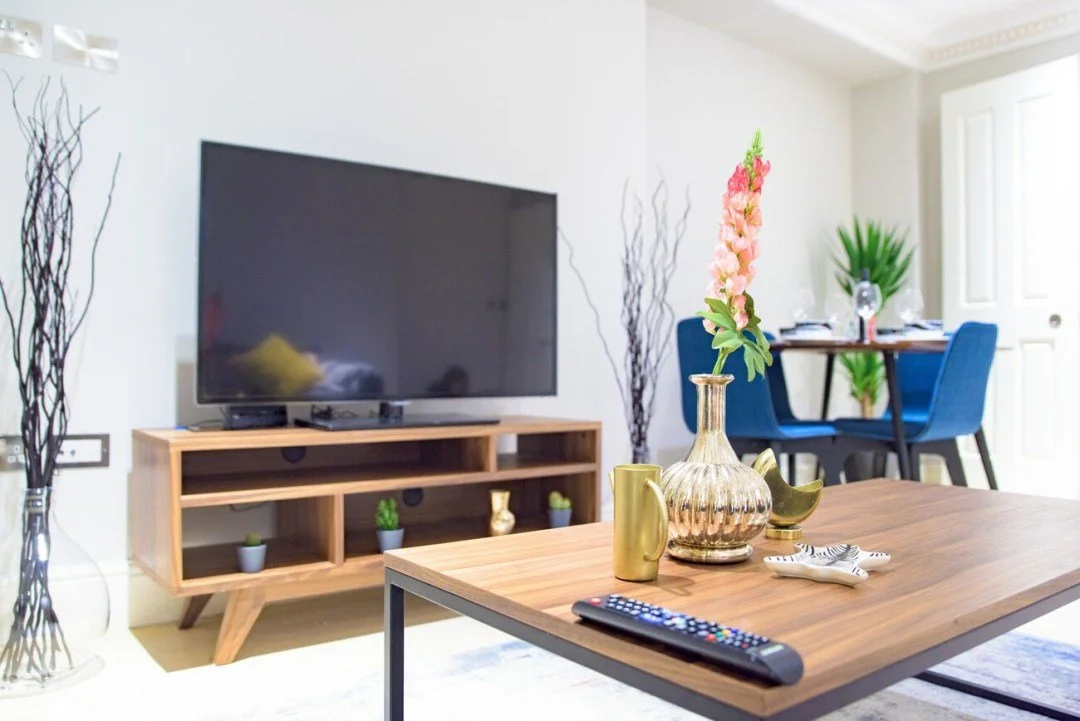 Living room with a wooden TV stand, a flat-screen TV, decorative vases, and a wooden coffee table with a remote and decorative items. In the background, there is a dining area with a table, blue chairs, and large windows.