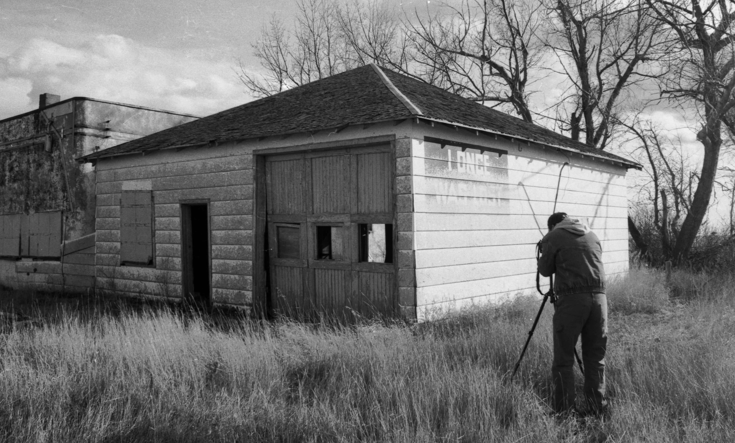 A person taking a photograph of an abandoned, weathered building with a sloped roof, broken windows, and overgrown grass in the foreground.