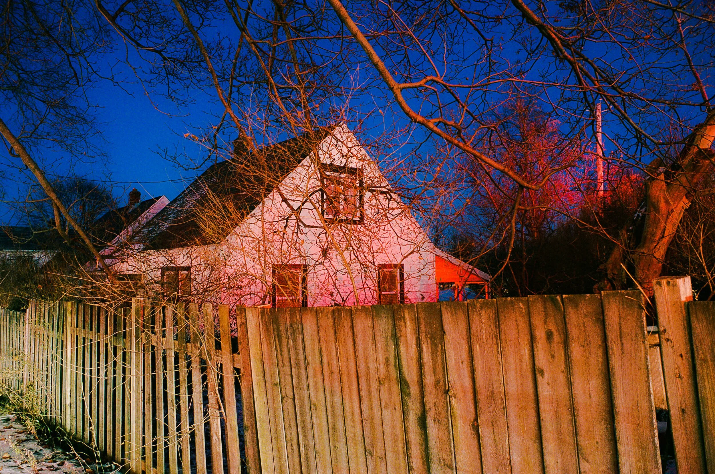 A house with a white exterior and black roof seen through leafless tree branches against a deep blue evening sky, with a wooden fence in the foreground.
