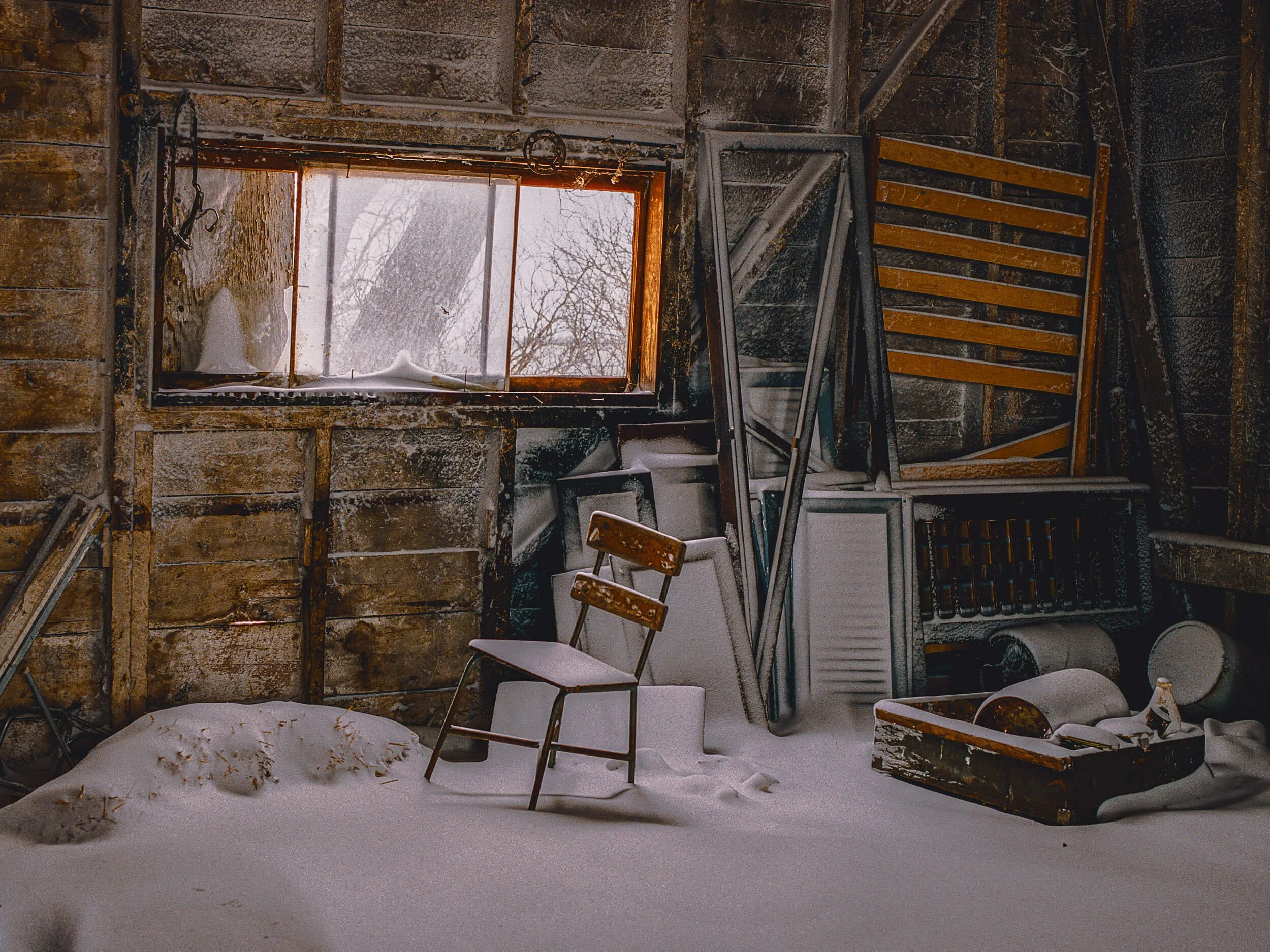 Snow-covered inside of a rustic, unfinished wooden shed or barn with a single window showing an outside snowy scene, filled with old furniture and tools covered in snow.