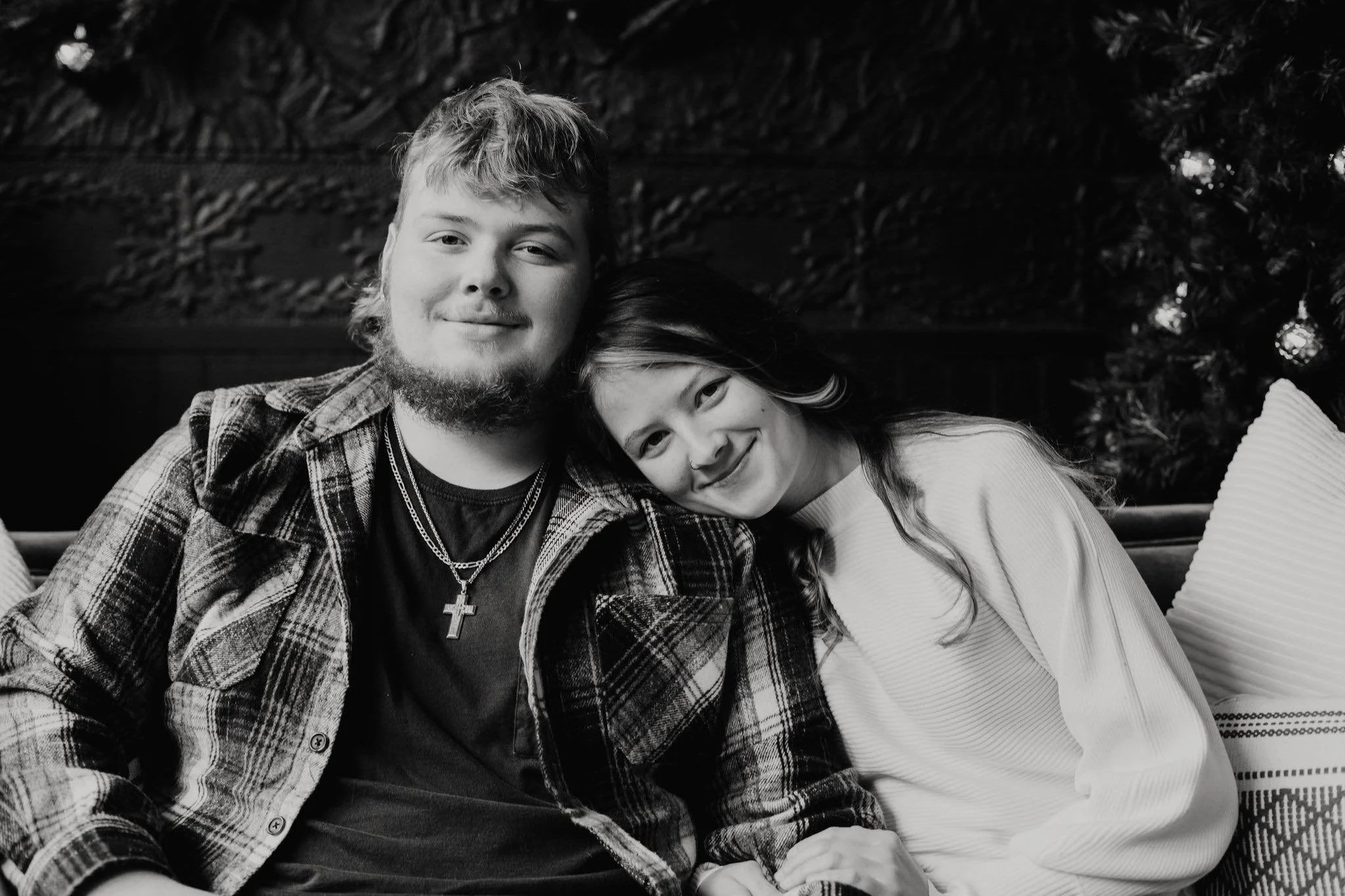 A young man with a beard and young woman with long hair sitting closely together, smiling, with a Christmas tree in the background.