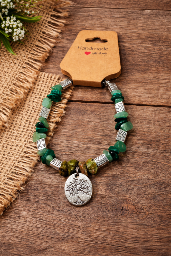 Green beaded necklace with a round silver pendant featuring a tree, displayed on a wooden surface beside burlap fabric and small white flowers.
