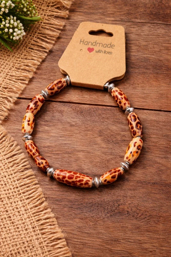 Beaded necklace with leopard print beads and silver spacers on a wooden surface, with a brown card that says 'Handmade with love' and a small bunch of white flowers on a burlap cloth.