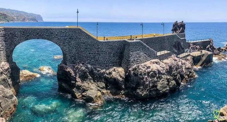 Stone bridge over rocky coastline with stairs leading down to water and a path along the top, overlooking the ocean.