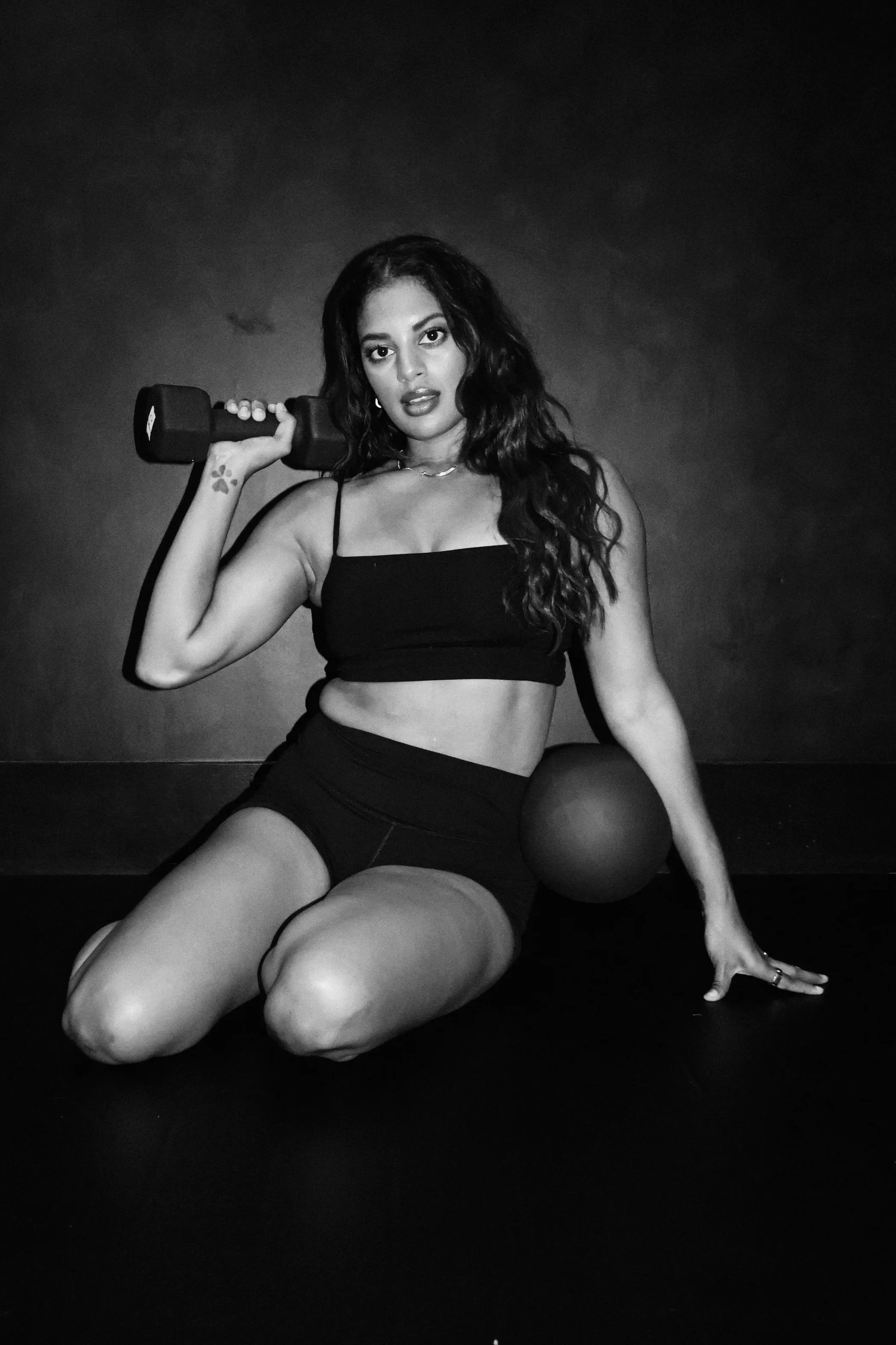 A woman with long, wavy hair and dark lipstick is kneeling on the floor, holding a dumbbell on her shoulder with her right hand, and resting her left hand on the ground. She is wearing a black sports bra and black shorts, and there is a medicine ball next to her. The background is plain and dark.