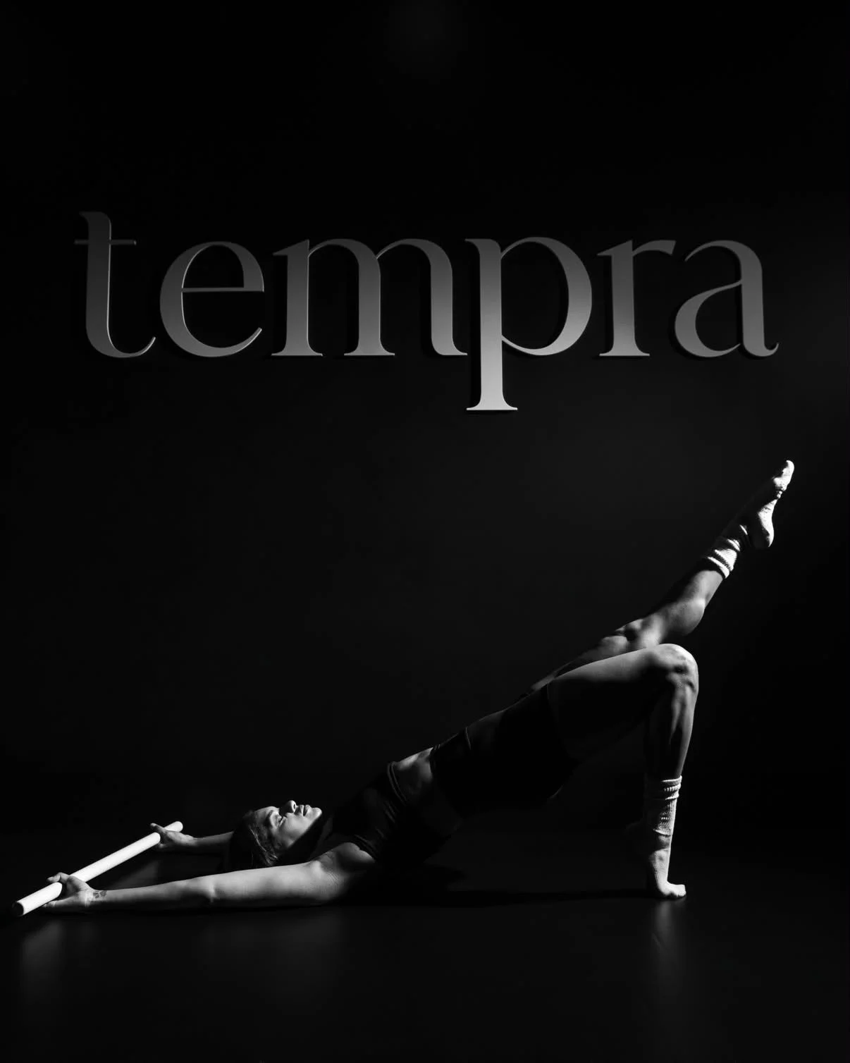 A woman exercising with a pole in a black and white studio, with the word 'tempra' displayed above in large letters.