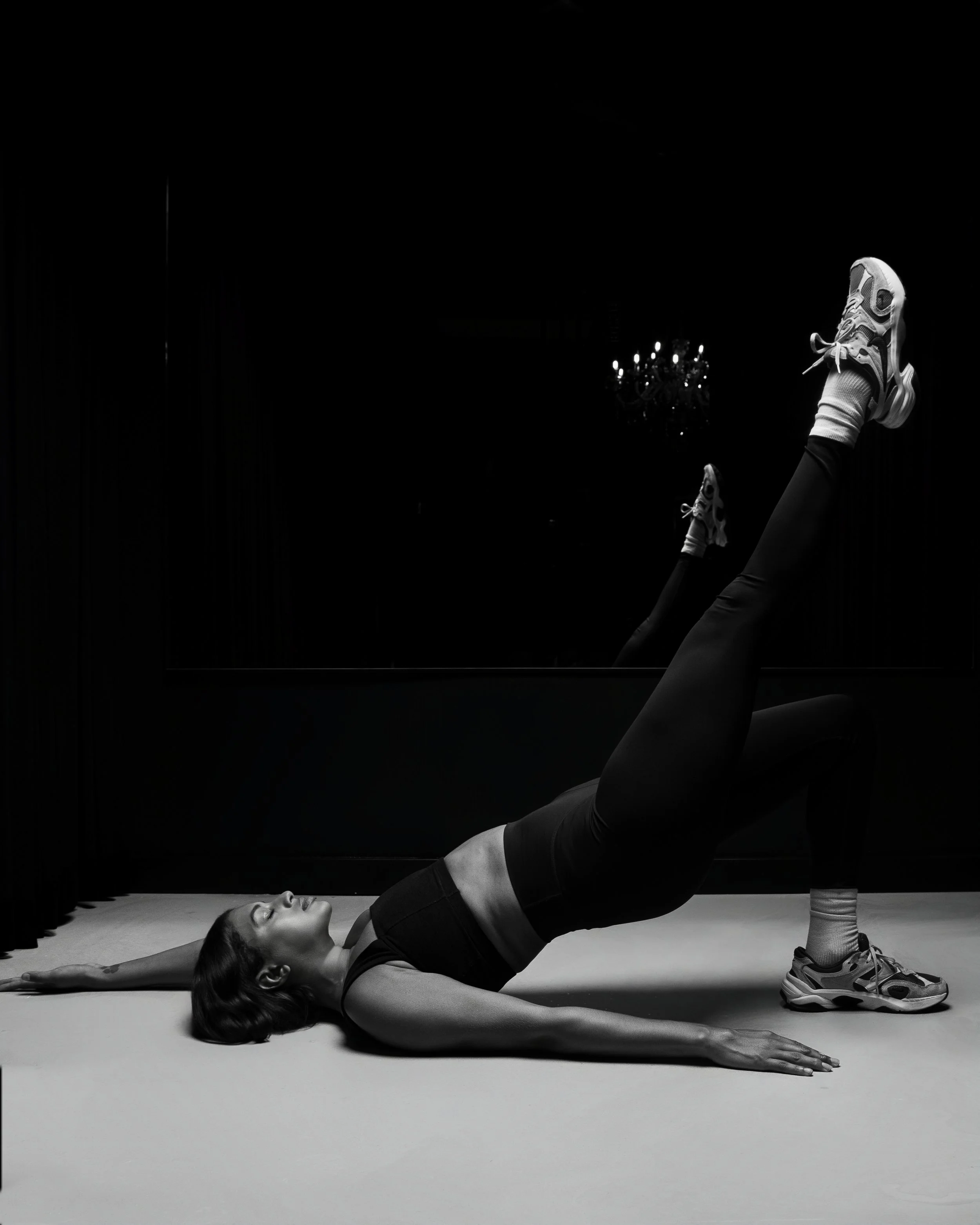 A woman performing a leg lift exercise on the floor, with one leg raised straight up and the other bent, in a dark room with a mirror and chandelier.