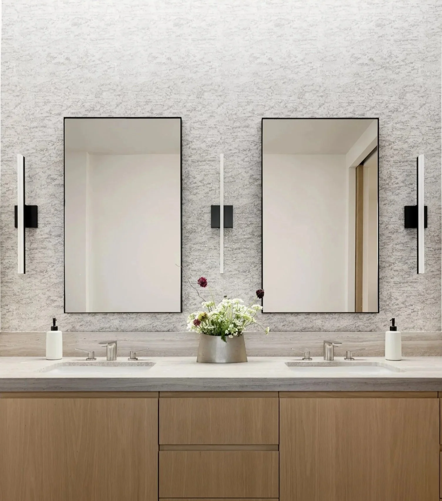Modern bathroom vanity with two mirrors, small flower arrangement, soap dispensers, and wall-mounted light fixtures.