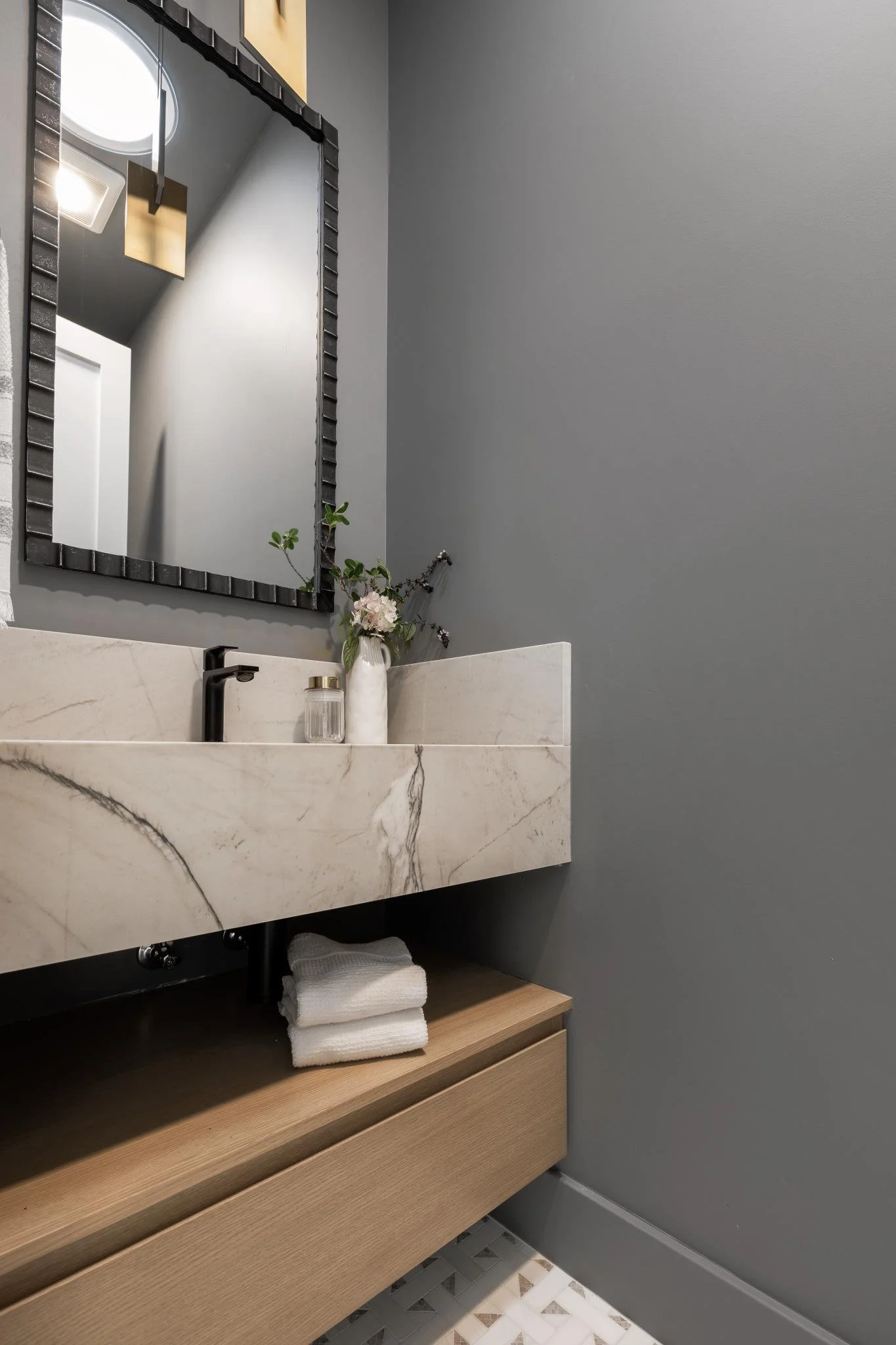 A modern bathroom with a marble sink, a framed mirror, and a small wooden cabinet below. There are folded white towels on the cabinet, a flower arrangement in a white vase, and a soap dispenser on the sink. The walls are painted gray.