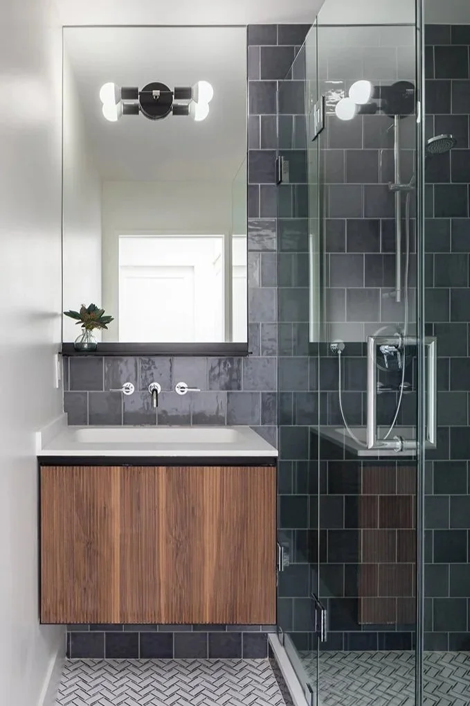 Modern bathroom with a gray tiled shower area, a small vanity with a wooden cabinet, white sink, and a large mirror above it. There's a unique light fixture on the wall and a small potted plant on the vanity.