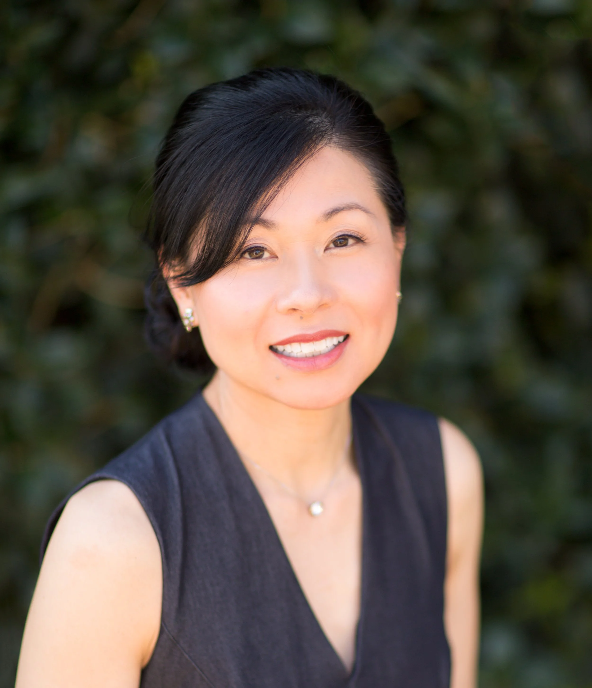 A woman with black hair wearing a black sleeveless top, pearl earrings, and a pearl necklace standing outdoors with greenery in the background.