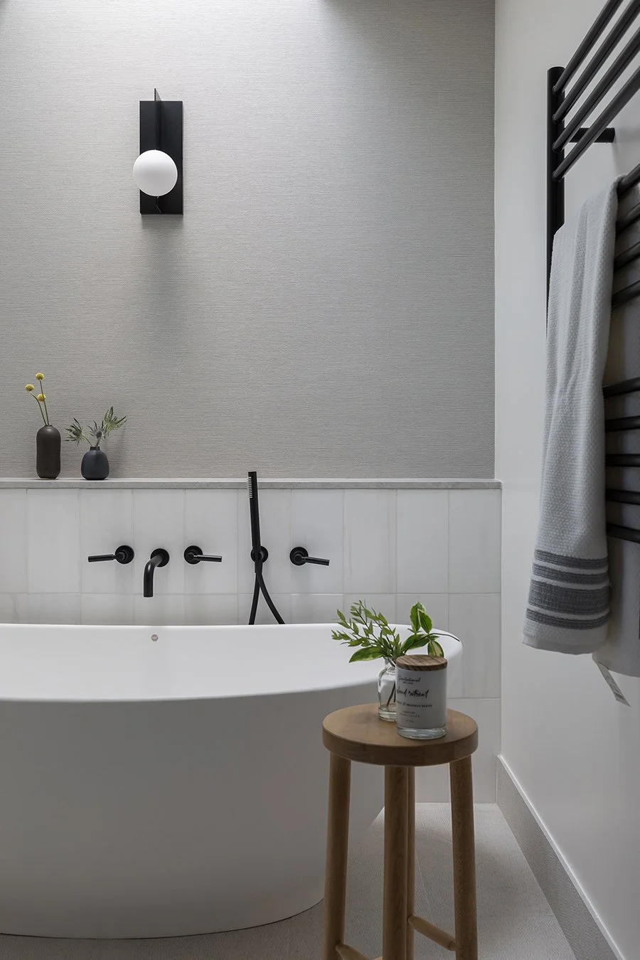 Minimalist bathroom with a white bathtub, black fixtures, a small wooden stool with a glass jar of green plants, a gray textured wall, black wall light, and a towel hanging on a black heated towel rack.