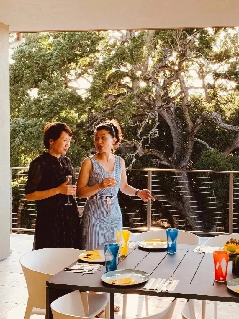 Two women having a conversation on a patio deck during daytime, with a large tree in the background, a black dining table with colorful glasses and plates.