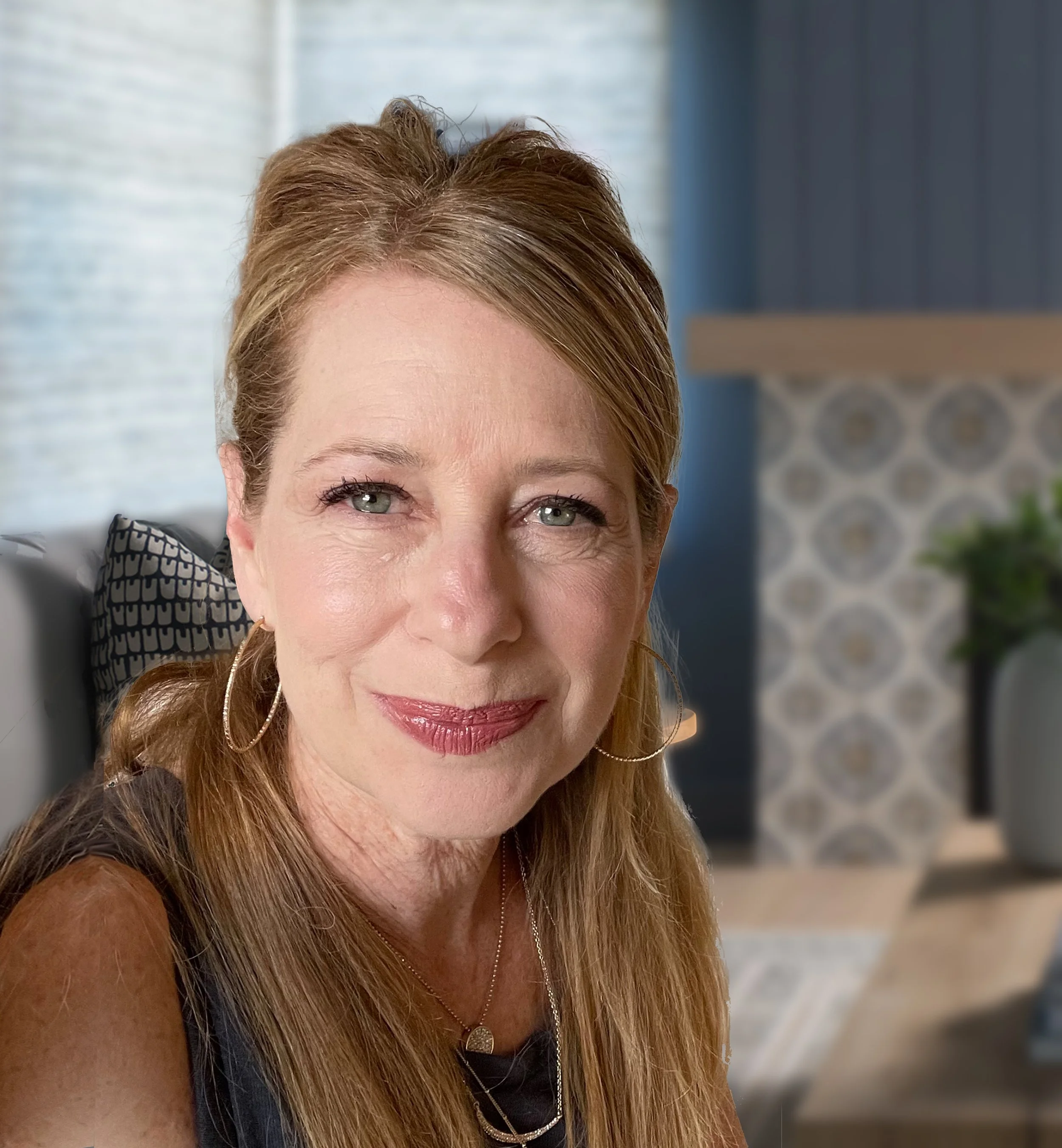 Close-up of a smiling middle-aged woman with long, light brown hair and hoop earrings in a modern, stylish living room.