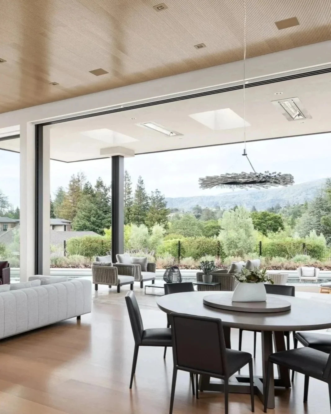 Modern living and dining area with large glass windows overlooking a lush green landscape and mountains, featuring a round dining table with black chairs, a white sofa, armchairs, and a decorative hanging light fixture.