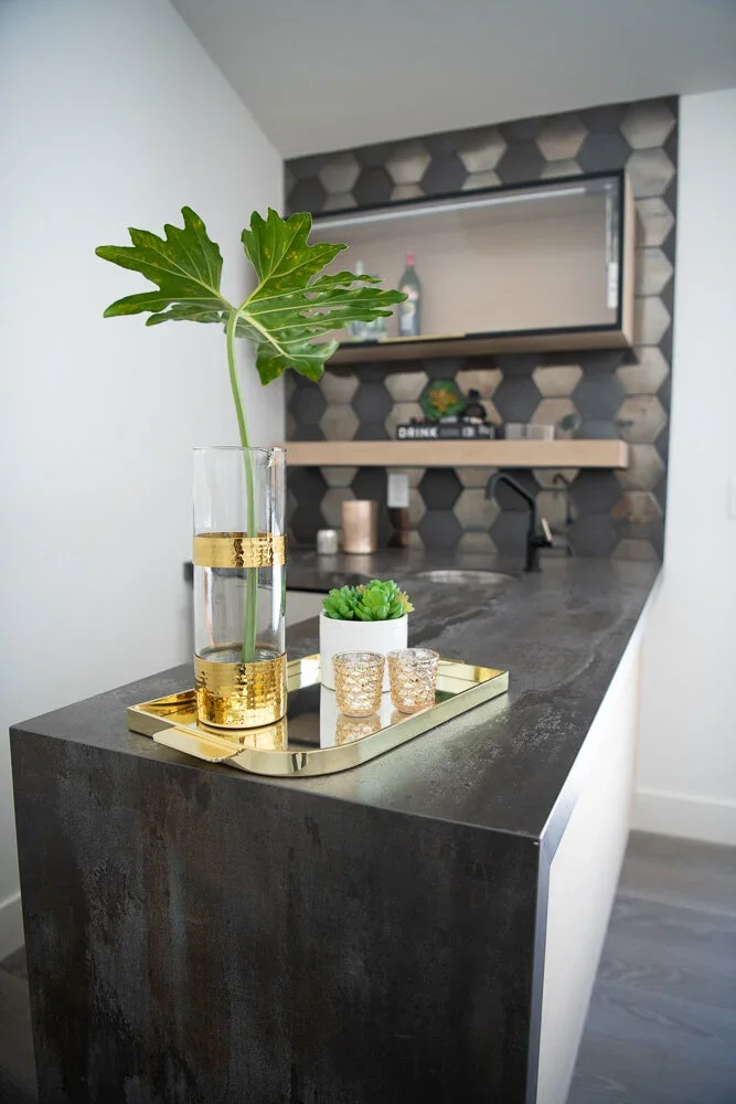 Modern kitchen countertop with a tall glass vase containing a large green leaf, a small potted succulent, and a gold tray with two textured glasses.