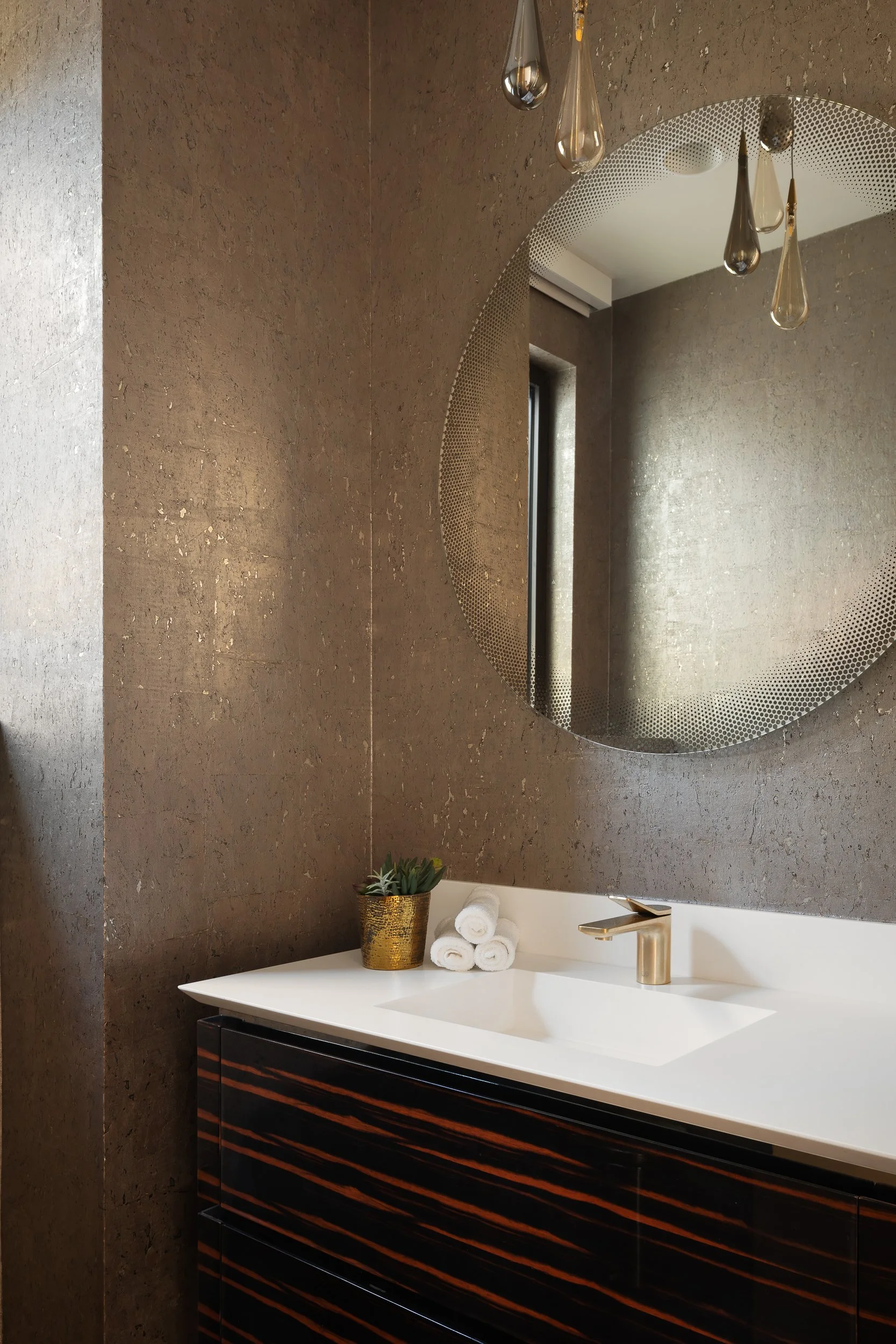 Modern bathroom with a round mirror, hanging teardrop-shaped pendant lights, a white countertop with a gold faucet, a small potted plant, and rolled white towels. The wall is textured with a dark, earthy tone.