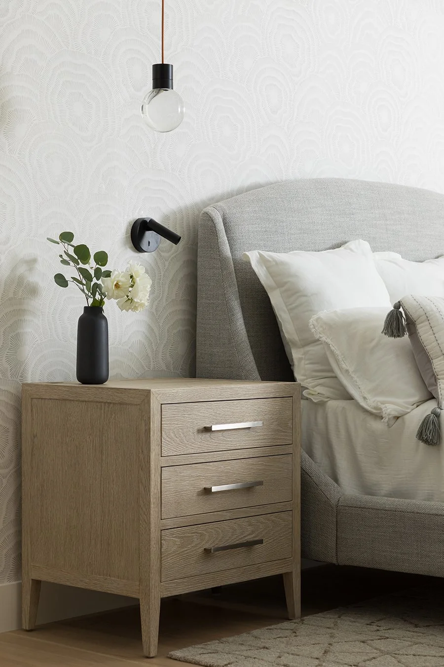 A bedroom with a light gray upholstered headboard, a wooden nightstand with three drawers, a black vase with white flowers on the nightstand, a black wall-mounted reading light, a wall-mounted black hanging light bulb, white bedding with decorative pillows, and a patterned beige rug.