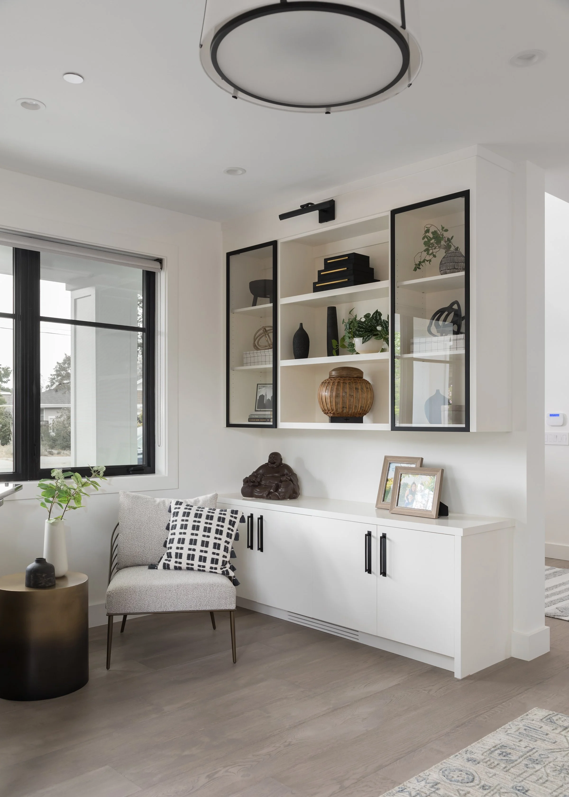 Living room with a white built-in cabinet, decorative items, and a gray armchair with a black and white patterned pillow, near a window with black framing.