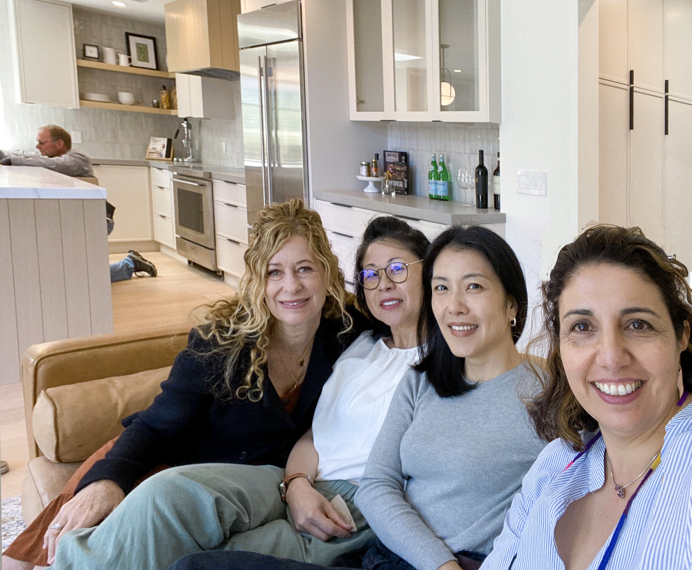 Four women sitting on a sofa in a modern kitchen, smiling at the camera, with a man in the background sitting at a table.