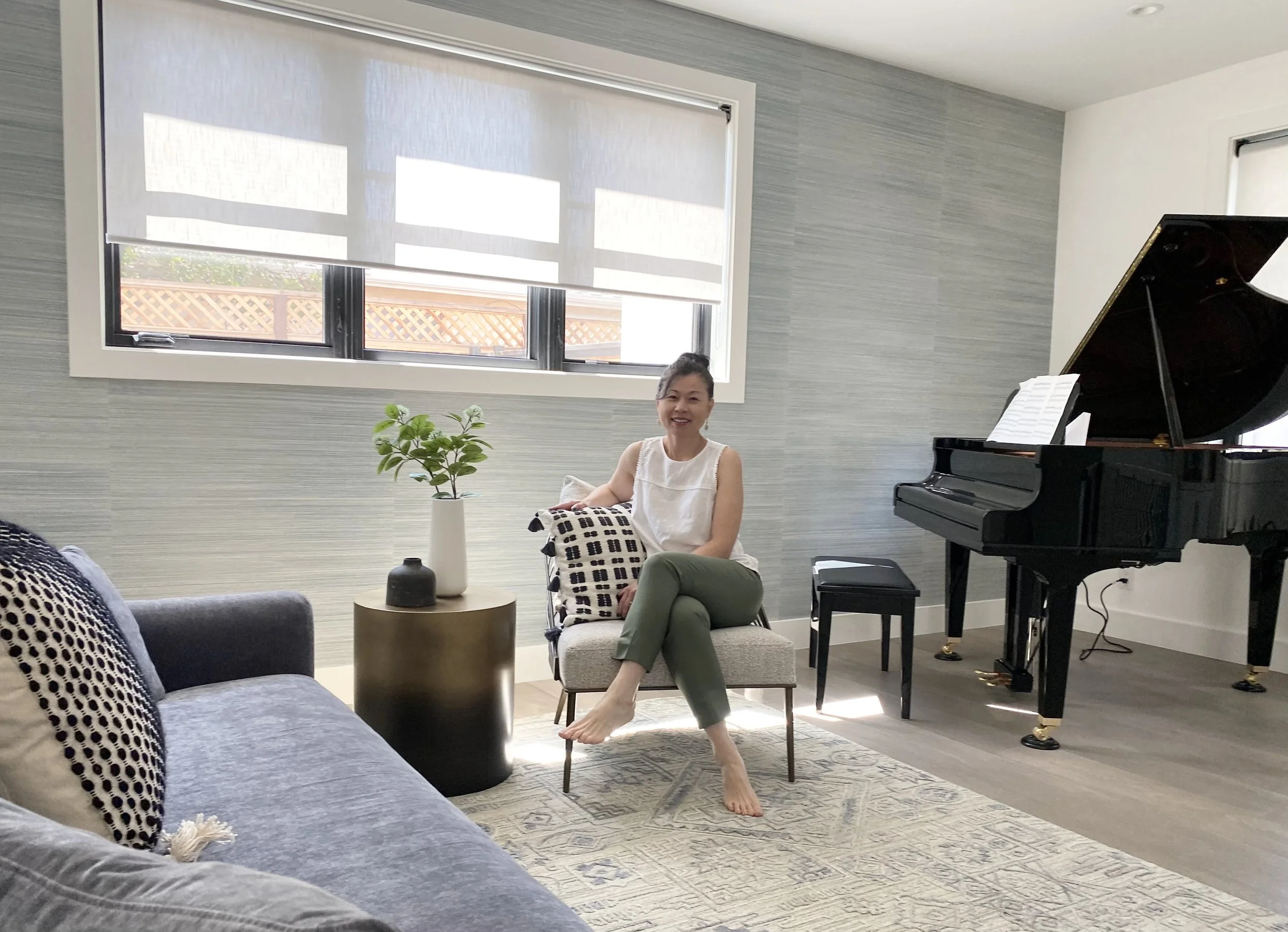 A woman sitting on a small chair in a bright living room, smiling at the camera. There is a black grand piano to her right, a window with blinds behind her, and a sofa with patterned pillows to her left. A small round side table with a green plant and a decorative object is in front of her, and a rug covers part of the wooden floor.