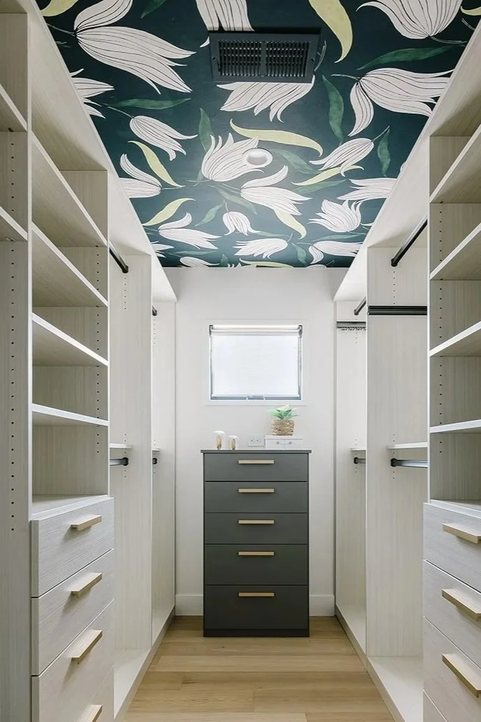 A walk-in closet with white shelves and drawers, a small dark gray dresser in the middle, a small window with a blind, a potted plant and toiletries on top, and a ceiling with a floral and leaf pattern.