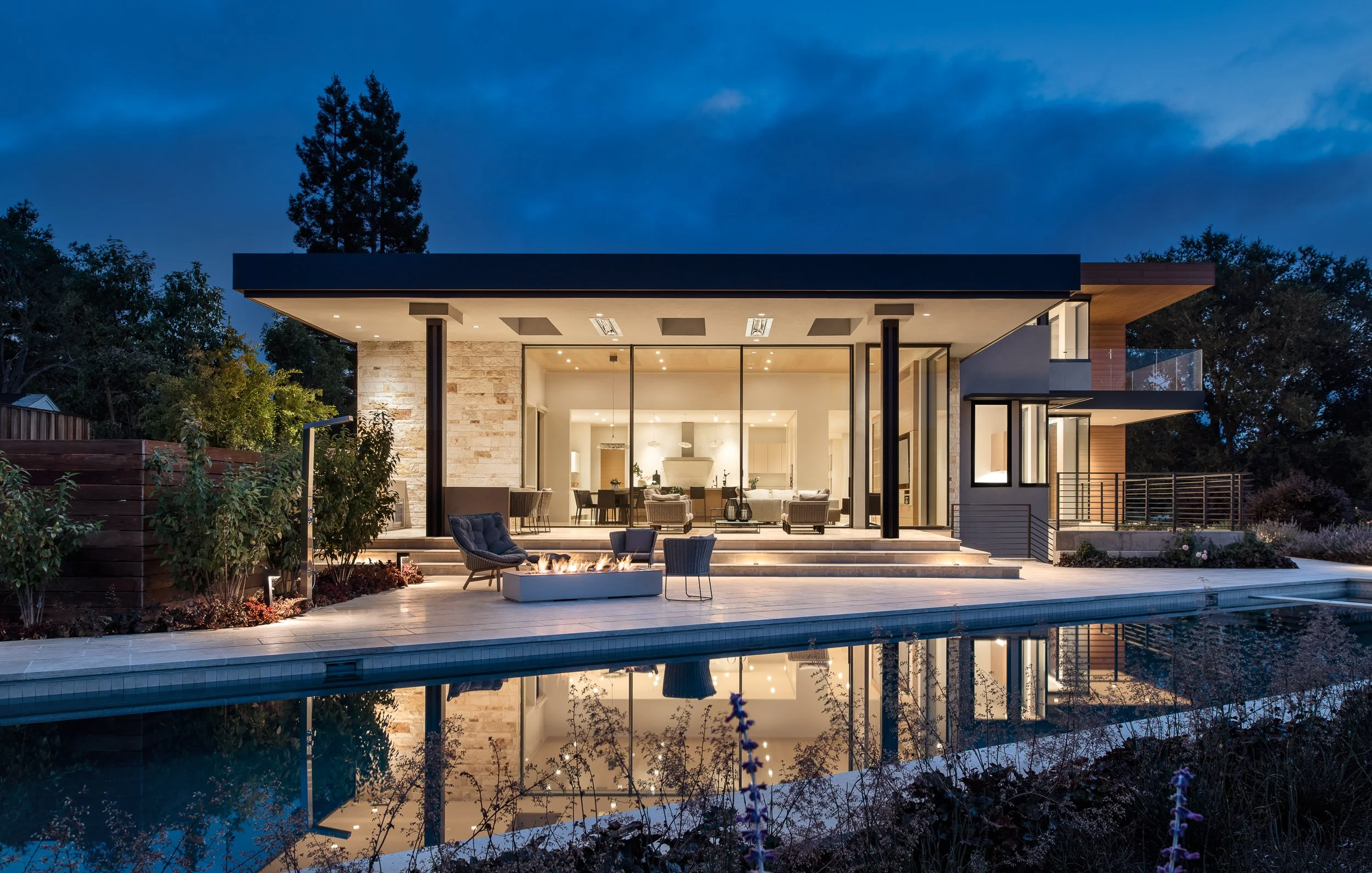 Modern house with large glass windows and a pool in the foreground, illuminated at dusk with outdoor seating and a firepit.