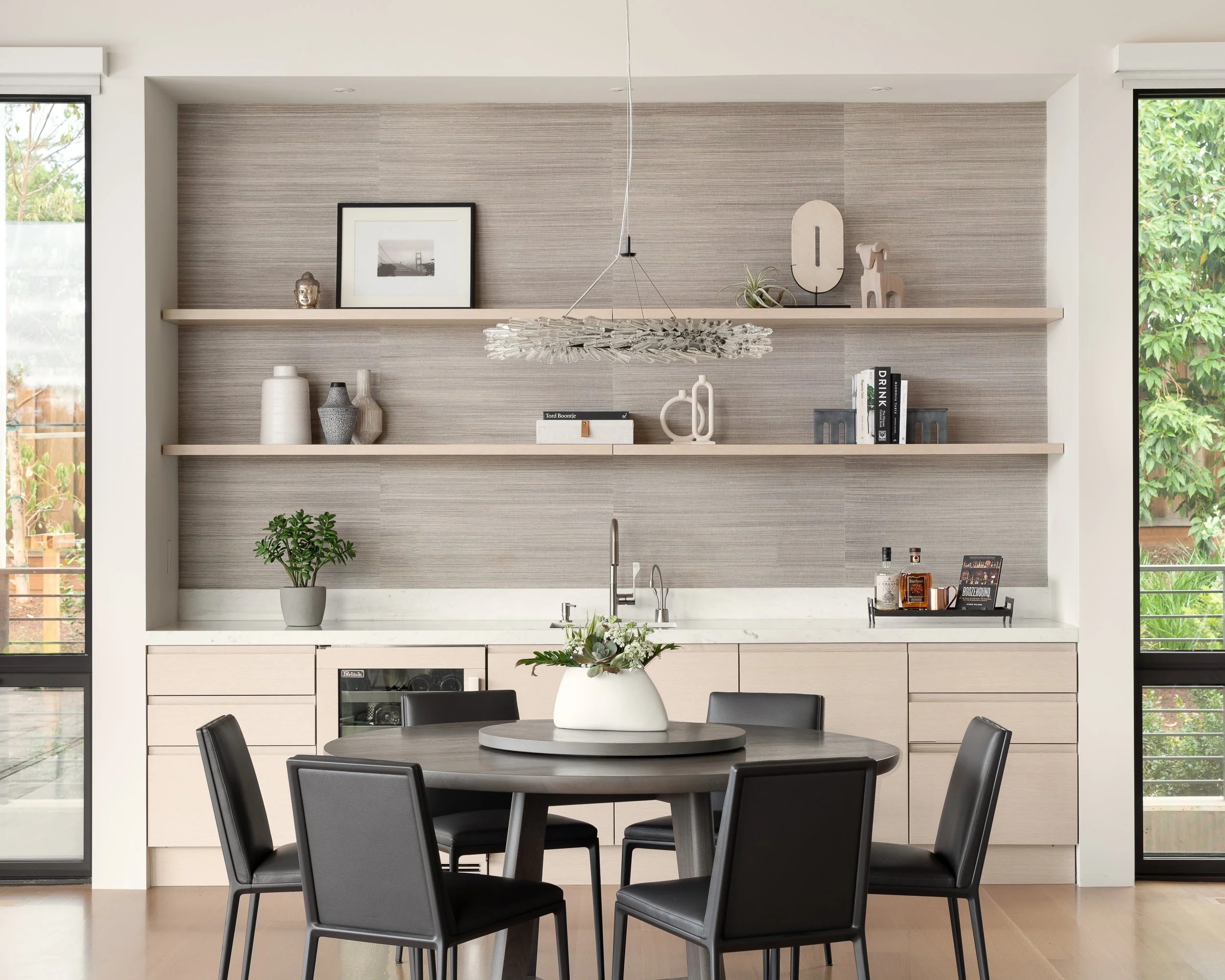 Modern kitchen with a round dining table, six black chairs, a built-in sink, open shelves with decorative vases, framed photo, and plants, with large windows showing greenery outside.