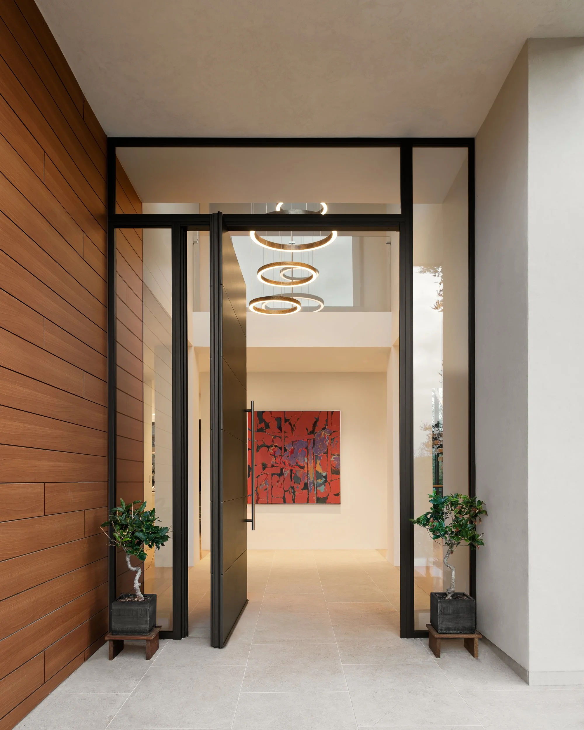 Modern entryway with wooden wall on the left, potted plants on either side of black open doors, and hanging circular light fixtures. A colorful abstract painting is visible inside.