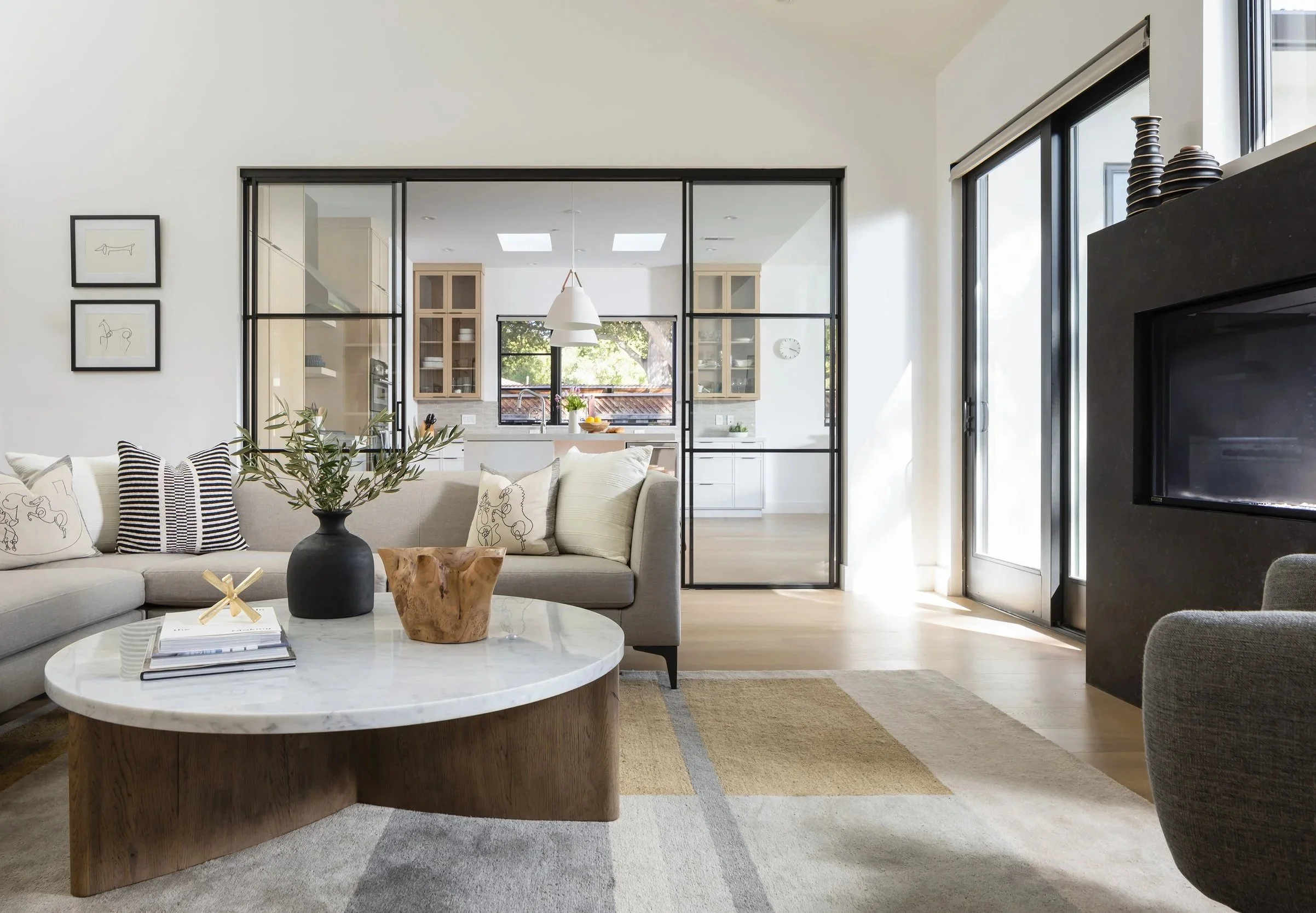 Modern living room with a white sectional sofa, decorative pillows, a marble-topped wooden coffee table with plants and books, large windows, and a view into a bright kitchen with cabinets, a hanging pendant light, and a clock on the wall.