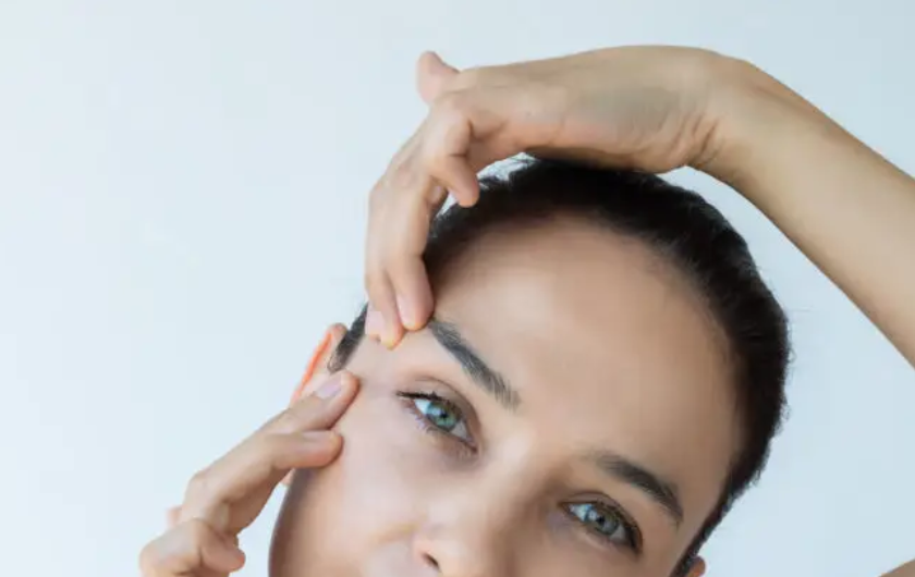 Close-up of a woman touching her forehead and face, with her hand on her hair and face, against a plain light background.