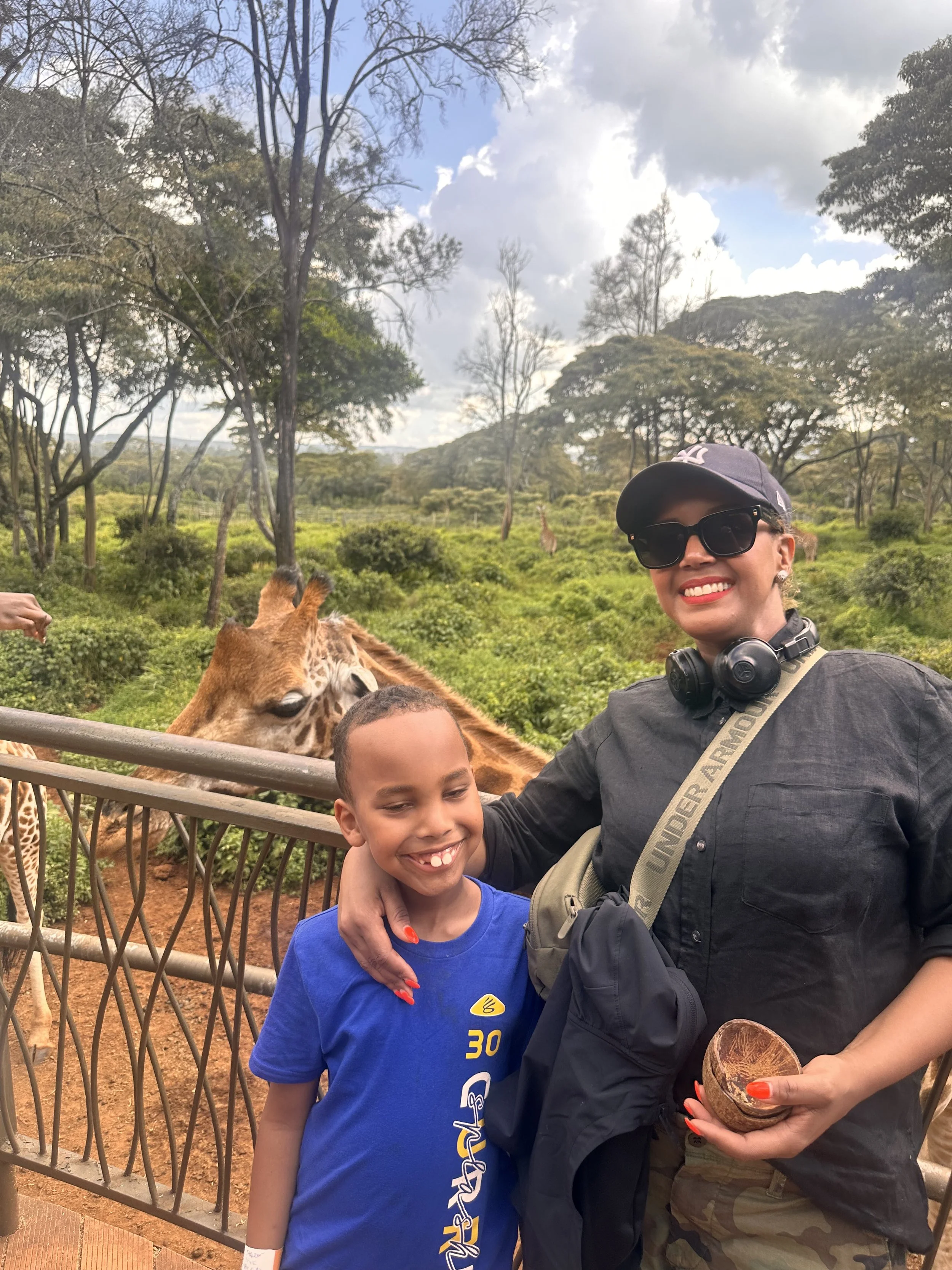 A woman and a boy smiling at the camera, standing near a fence with giraffes in the background at a wildlife park or zoo.