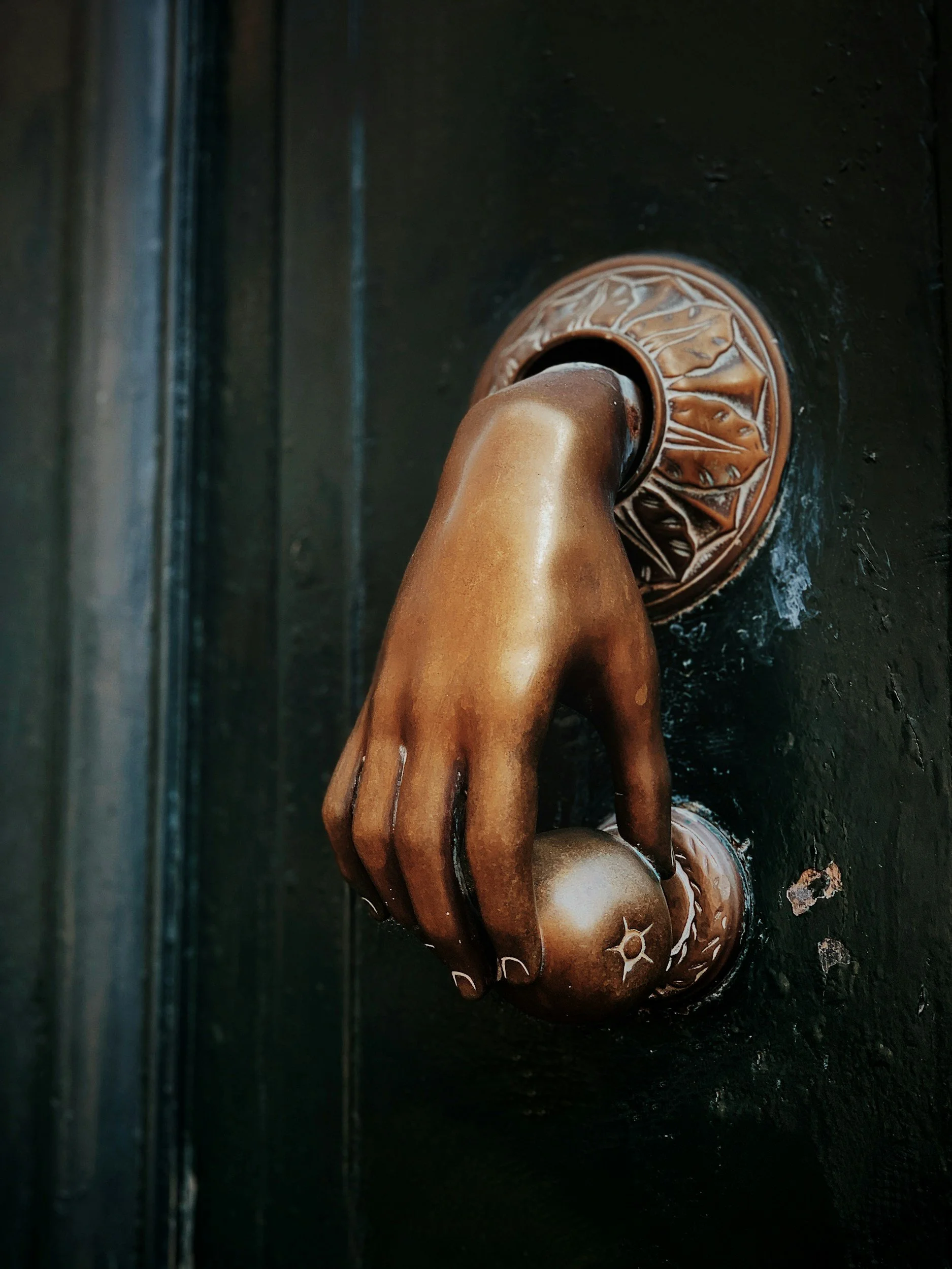 Close-up of a hand door knocker, bronze-colored hand sculpture with detailed fingers, resting on a dark surface with decorative round ornaments nearby.