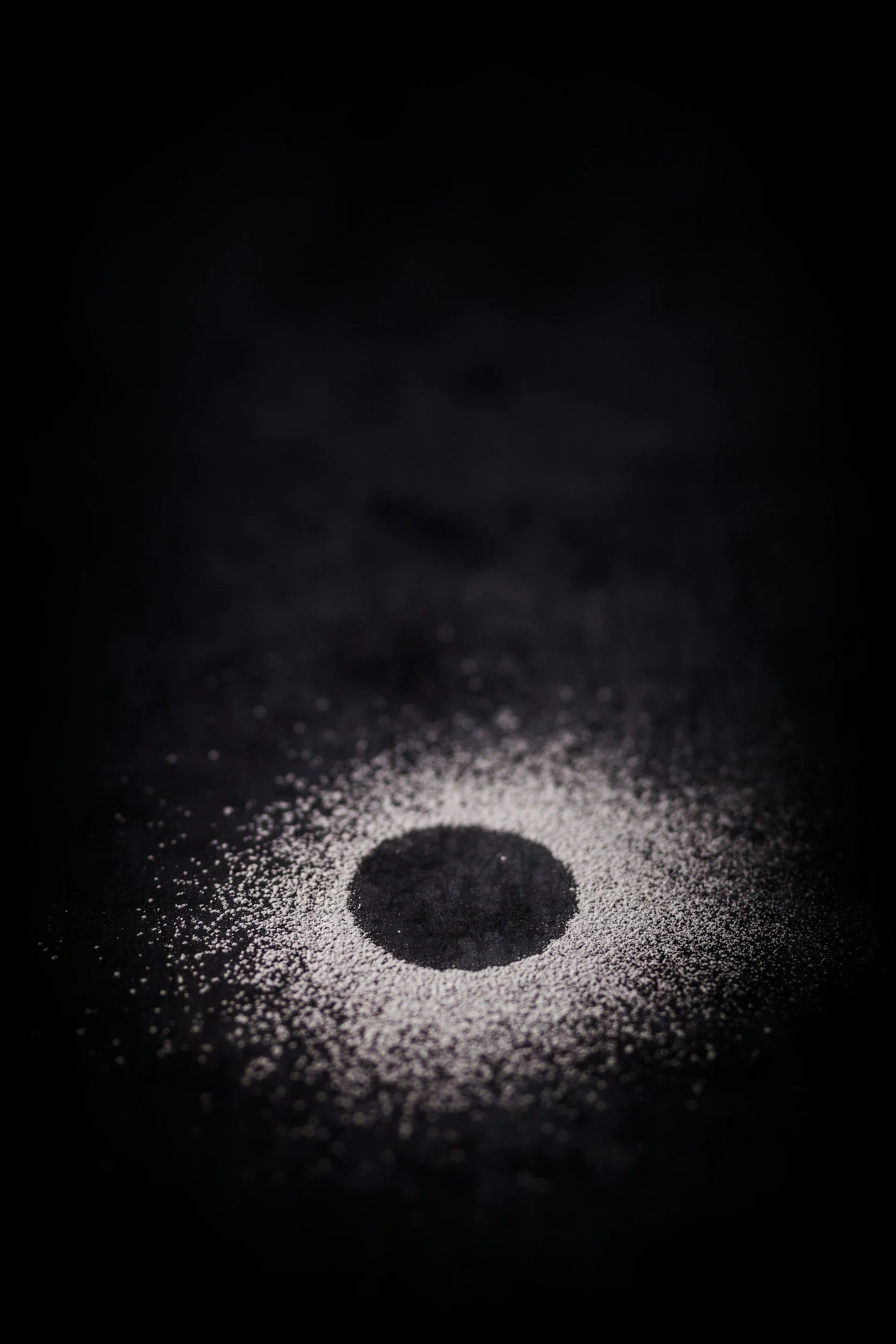 Black surface with a circular pattern of salt, in the center, with a small dark circle within it, and a dark background surrounding the scene.