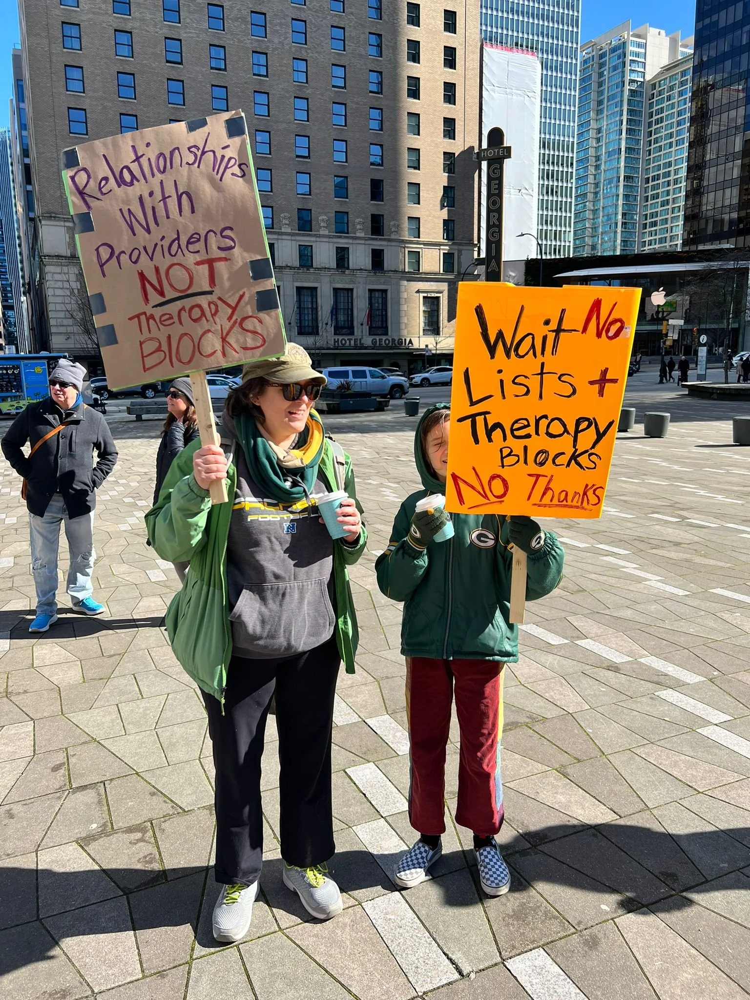 Two people holding protest signs in an urban area, surrounded by a few other individuals. One woman's sign reads, 'Relationships with Providers NOT Therapy BLOCKS,' and the child’s sign states, 'Wait No Lists + Therapy Blocks No Thanks.'