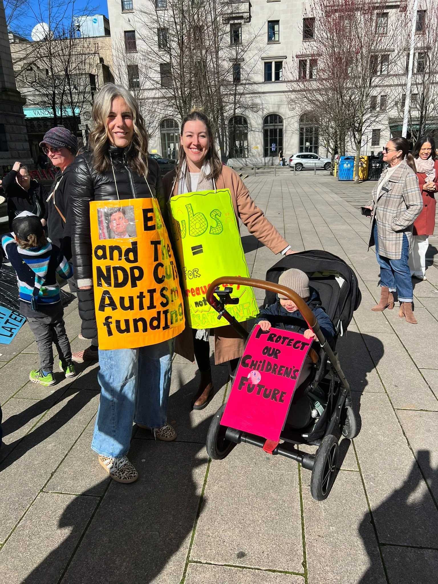 Two women at a protest holding colorful signs. One sign promotes autism funding, the other advocates for protecting children's future. A child in a stroller is also present.
