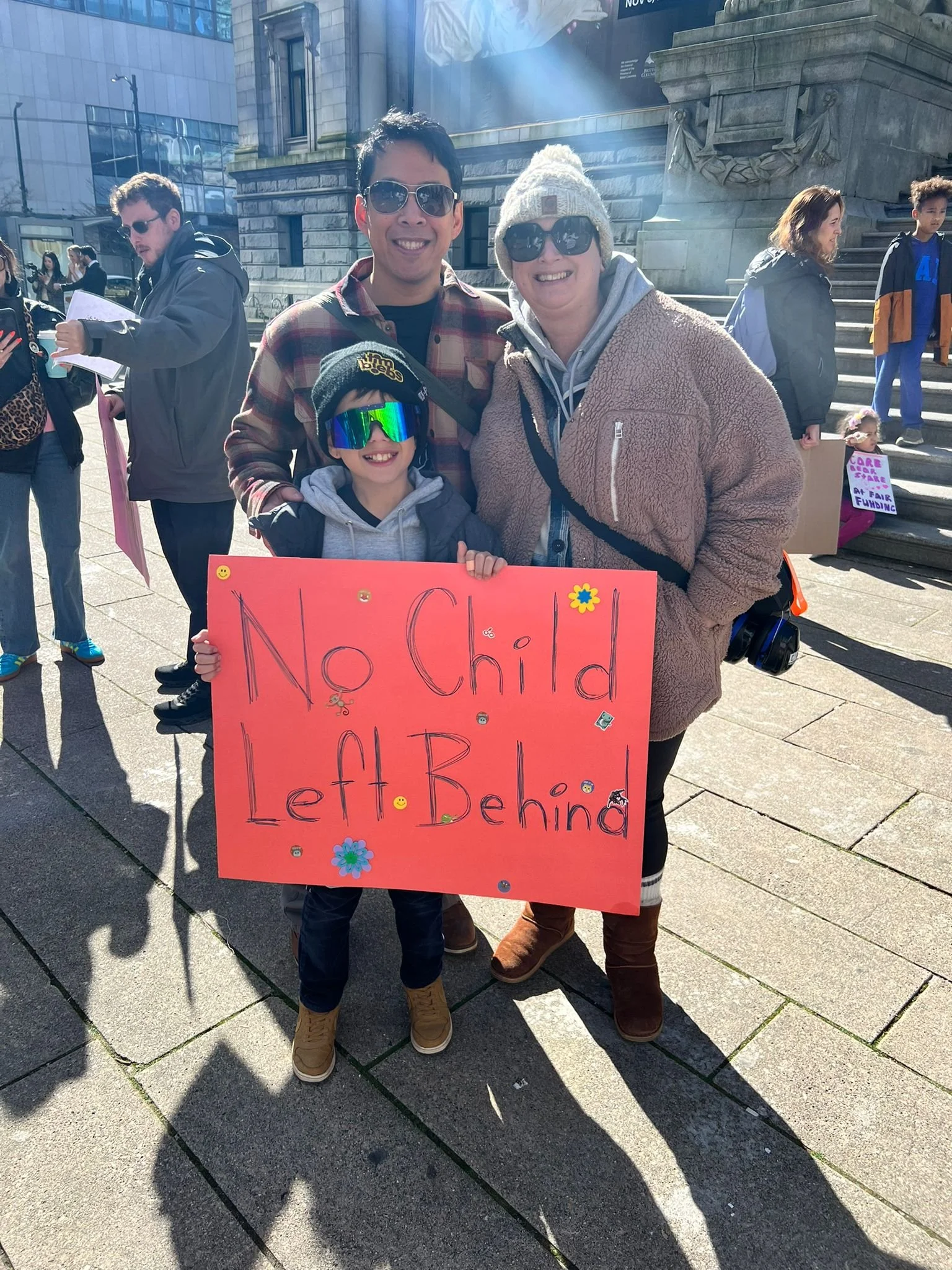 A child holding a pink protest sign that reads 'No Child Left Behind,' standing with two adults during a rally or protest on a city street. The child is wearing sunglasses and a beanie, and the woman beside them is wearing sunglasses and a beige jacket. The protest scene includes other people in the background, some holding signs.