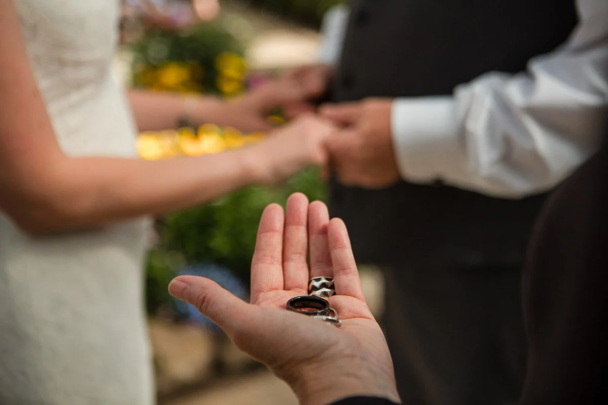Hand holding wedding rings during wedding ceremony while couple hold hands in the background.